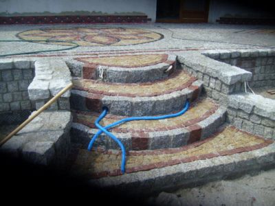 Schody zewnętrzne wyłożone mozaiką z kamienia naturalnego w odcieniach szarości, beżu i czerwieni, z widocznymi rurami instalacji elektrycznej i fragmentem nawierzchni z dekoracyjnym wzorem.